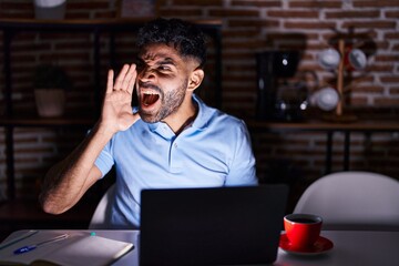 Hispanic man with beard using laptop at night shouting and screaming loud to side with hand on mouth. communication concept.