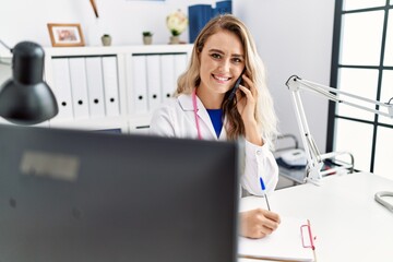 Young woman wearing doctor uniform writing on document talking on smartphone at clinic