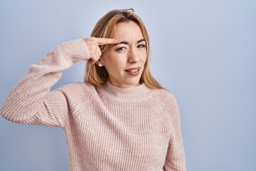 Hispanic woman standing over blue background pointing unhappy to pimple on forehead, ugly infection...