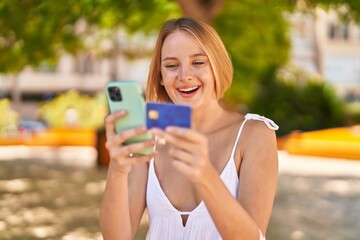 Young blonde woman using smartphone and credit card at park