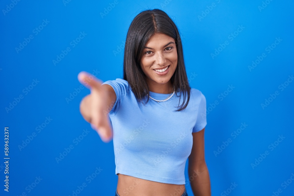 Sticker Brunette young woman standing over blue background smiling friendly offering handshake as greeting and welcoming. successful business.