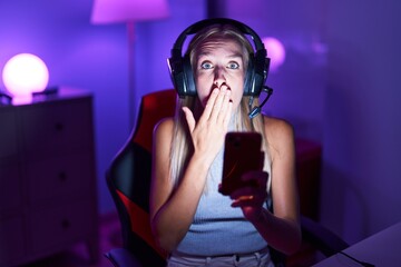 Young caucasian woman playing video games with smartphone covering mouth with hand, shocked and afraid for mistake. surprised expression