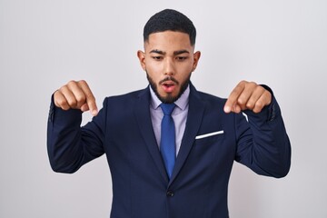 Young hispanic man wearing business suit and tie pointing down with fingers showing advertisement, surprised face and open mouth