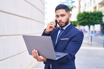 Young latin man business worker using laptop talking on smartphone at street