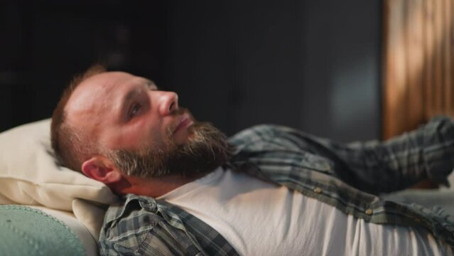 Bearded man takes comfortable pose on sofa to take rest and sleep. Male with bald patches having cardiac disease relaxes in living room at home close view