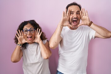 Middle age hispanic couple together over pink background smiling cheerful playing peek a boo with hands showing face. surprised and exited