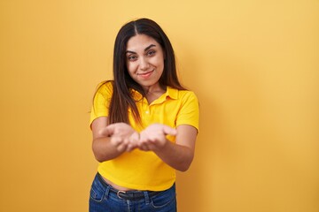 Young arab woman standing over yellow background smiling with hands palms together receiving or giving gesture. hold and protection