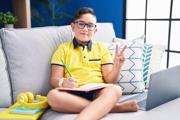 Young hispanic kid doing homework sitting on the sofa smiling with happy face winking at the camera...
