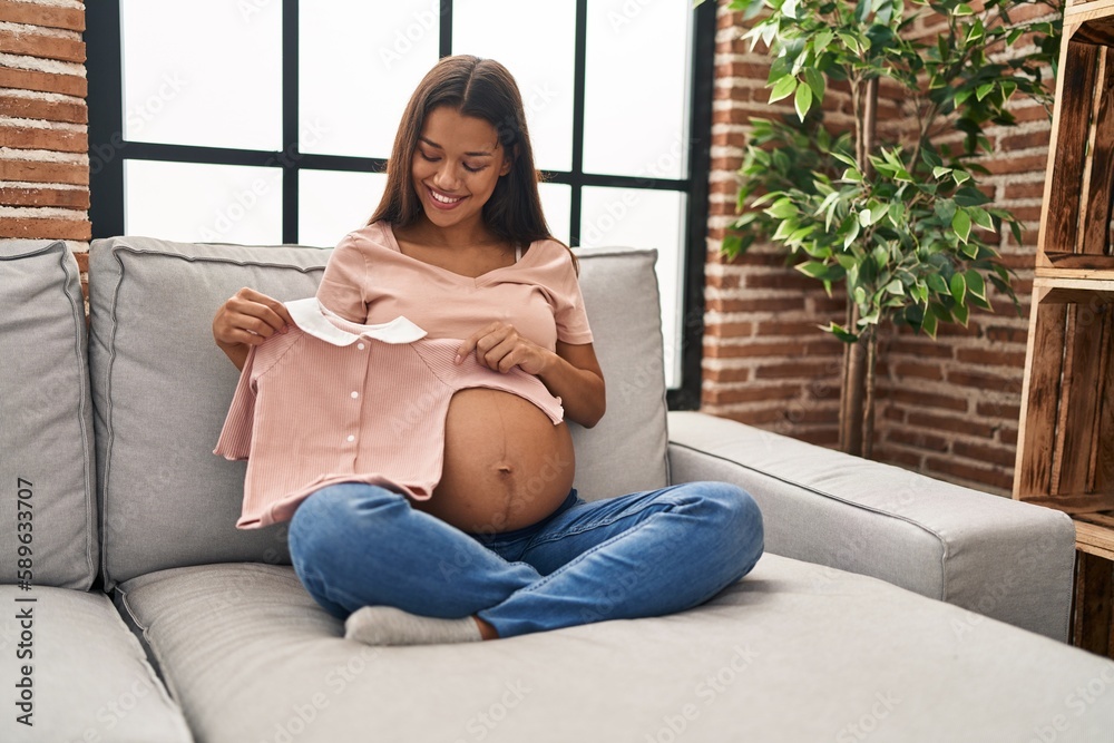 Sticker young latin woman pregnant holding baby clothes at home