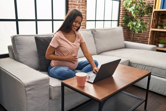 Young latin woman pregnant using laptop drinking coffee at home
