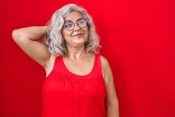 Middle age woman with grey hair standing over red background smiling confident touching hair with...