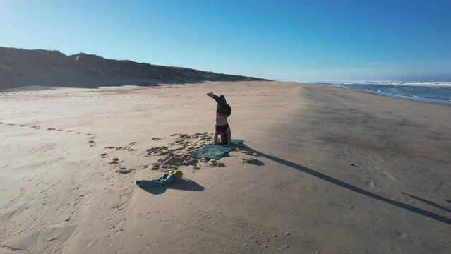 A gorgeous woman does hatha yoga postures on her floor mat alone at the beach with the drone making a circle around her, then the floor mat comes back on her head with a gust of wind