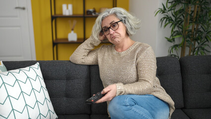 Middle age woman with grey hair watching tv sitting on sofa with boring expression at home