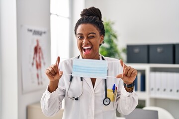 Young african american with braids wearing doctor uniform holding safety mask winking looking at...
