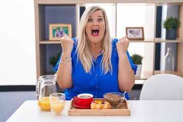 Obraz na płótnie Canvas Caucasian plus size woman eating breakfast at home celebrating surprised and amazed for success with arms raised and open eyes. winner concept.