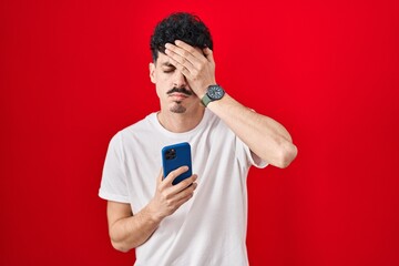 Hispanic man using smartphone over red background yawning tired covering half face, eye and mouth with hand. face hurts in pain.