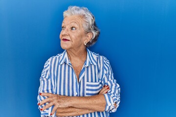 Senior woman with grey hair standing over blue background smiling looking to the side and staring away thinking.