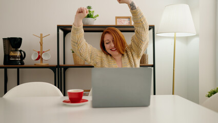 Young redhead woman using laptop with celebrate expression at home