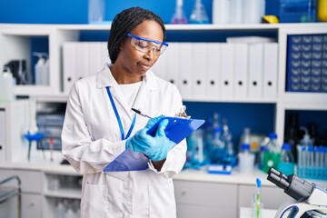 African american woman wearing scientist uniform writing on clipboard laboratory