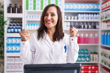 Middle age brunette woman working at pharmacy drugstore celebrating surprised and amazed for success with arms raised and open eyes. winner concept.