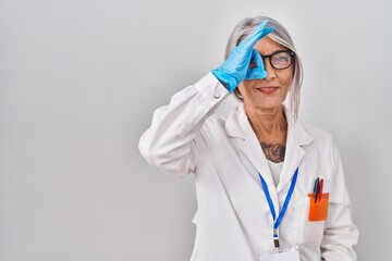 Middle age woman with grey hair wearing scientist robe doing ok gesture with hand smiling, eye looking through fingers with happy face.