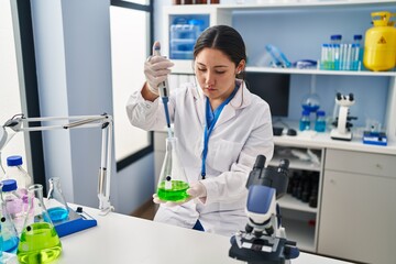Fototapeta na wymiar Young hispanic woman wearing scientist uniform using pipette working at laboratory