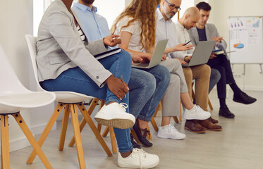 Young business people sitting on the chairs in a row with resumes and laptops in their hands chatting and talking with each other. Job candidates seekers waiting for interview invitation turn.