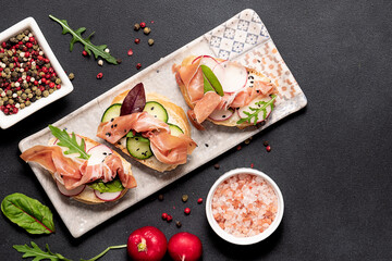 Set of bruschetta with jamon or prosciutto, vegetables and fresh arugula leaves on the ceramic serving board on black stone background top view. Healthy homemade breakfast or lunch idea.