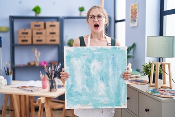 Young caucasian woman holding canvas at art studio angry and mad screaming frustrated and furious, shouting with anger. rage and aggressive concept.
