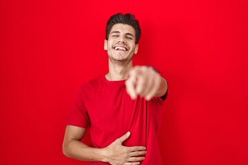 Young hispanic man standing over red background laughing at you, pointing finger to the camera with...