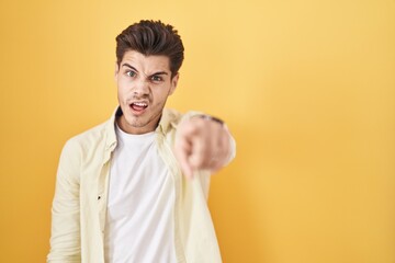 Young hispanic man standing over yellow background pointing displeased and frustrated to the...