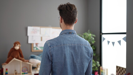 Young hispanic man preschool teacher standing on back view at kindergarten