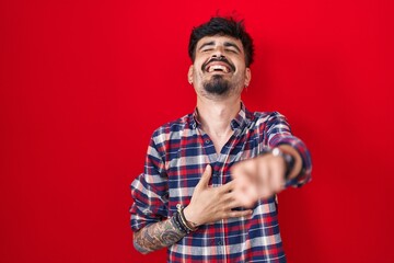 Young hispanic man with beard standing over red background laughing at you, pointing finger to the camera with hand over body, shame expression