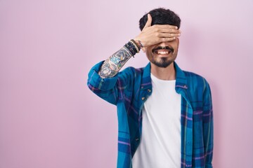 Young hispanic man with beard standing over pink background smiling and laughing with hand on face covering eyes for surprise. blind concept.