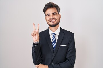 Young hispanic man with tattoos wearing business suit and tie smiling with happy face winking at...