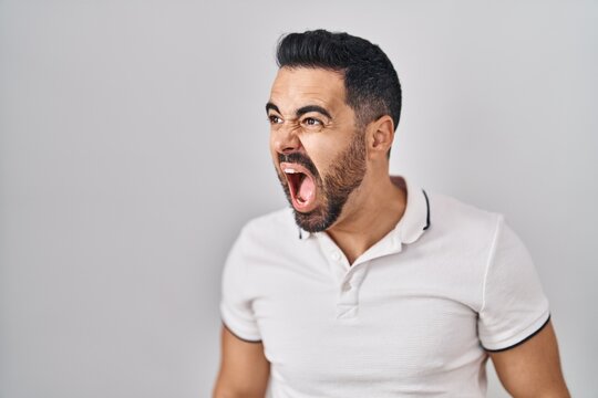 Young hispanic man with beard wearing casual clothes over white background angry and mad screaming frustrated and furious, shouting with anger. rage and aggressive concept.