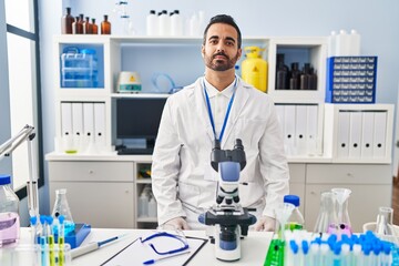 Fototapeta premium Young hispanic man with beard working at scientist laboratory relaxed with serious expression on face. simple and natural looking at the camera.
