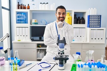 Young hispanic man with beard working at scientist laboratory looking confident at the camera smiling with crossed arms and hand raised on chin. thinking positive.