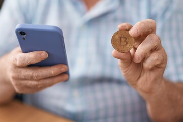 Middle age grey-haired man using smartphone holding bitcoin at office