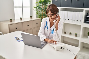 Young beautiful hispanic woman doctor using laptop talking on telephone at clinic