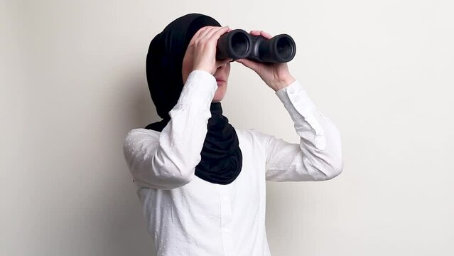 Young woman in hijab looking through binoculars on white background.