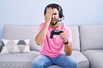 Hispanic young man playing video game holding controller sitting on the sofa yawning tired covering half face, eye and mouth with hand. face hurts in pain.