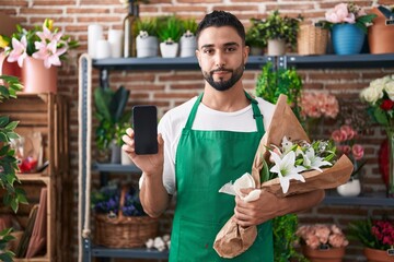 Hispanic young man working at florist shop showing smartphone screen relaxed with serious expression on face. simple and natural looking at the camera.