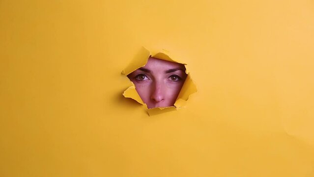 Young woman looking through hole on yellow background.