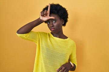 African young woman standing over yellow studio making fun of people with fingers on forehead doing...
