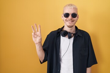 Young caucasian man wearing sunglasses standing over yellow background showing and pointing up with fingers number three while smiling confident and happy.