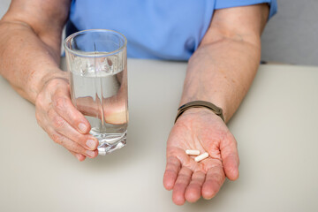 Senior woman sitting at the table and holding in the old wrinkled hands white capsules, vitamins or pills for treatment and water glass, healthcare and medicine concept
