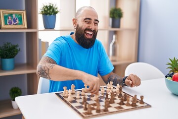 Young bald man playing chess sitting on table at home