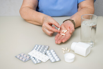 Senior woman sitting at the table and holding in the old wrinkled hands white capsules, vitamins or pills for treatment, healthcare and medicine concept