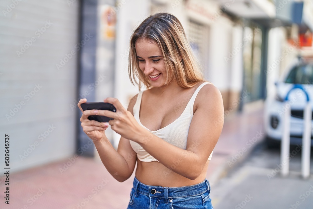 Poster young blonde woman smiling confident playing video game at street